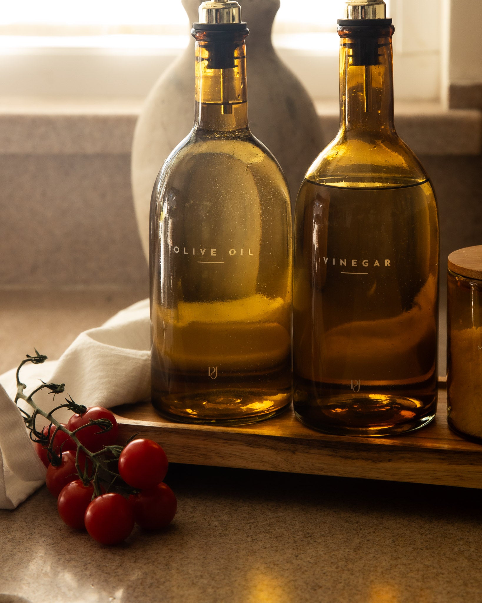 Two bottles of olive oil and vinegar on a wooden tray with tomatoes and a jar in the background.