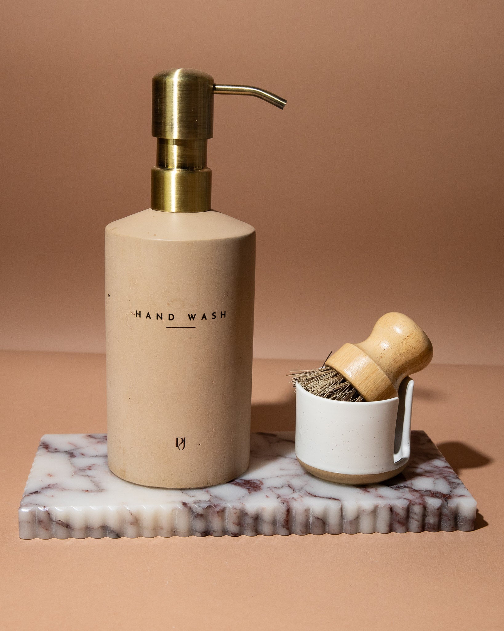 Hand wash bottle with gold pump and wooden brush on marble tray against a brown background