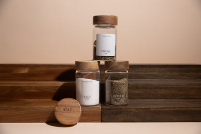Three glass jars with wooden lids on a wooden surface, featuring labels for 'Thyme', 'Salt', and 'Pepper'.