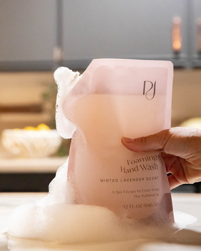 Hand holding a pink foaming hand wash bottle with visible brand logo, set against a blurred indoor background.