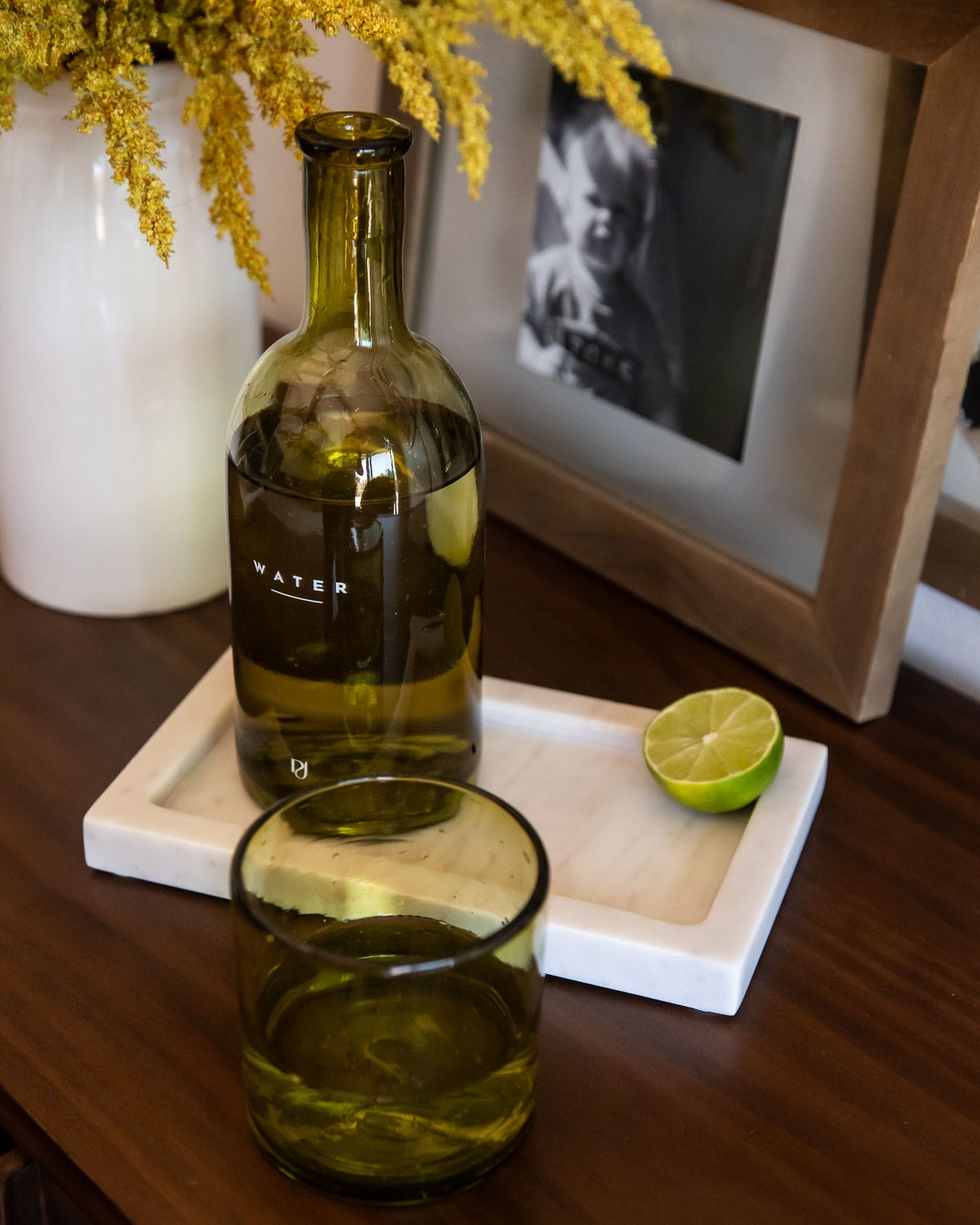 Green glass bottle labeled 'WATER' on a wooden surface with a lime and photo frame in the background.