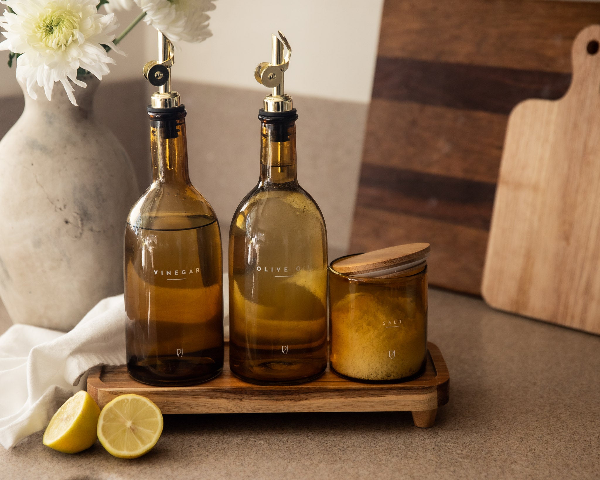 Two olive oil bottles with nozzles on a wooden tray with lemons and a vase in the background.