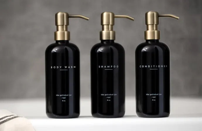 Three black refillable spa bottles labeled body wash, shampoo, and conditioner with gold pumps.